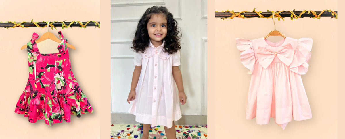 Smiling girl in a light pink dress stands between two kids' dresses, floral and ruffled, on hangers